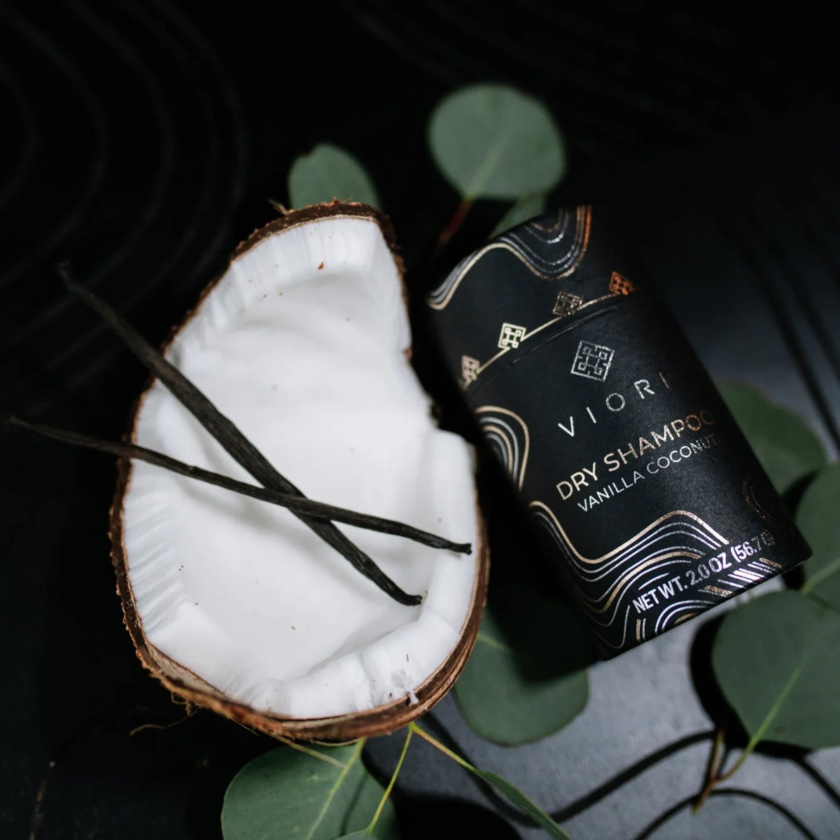 Viori tørshampoo med vanilje og kokos i sort emballage, omgivet af kokosnød og eucalyptusblade, til sund hårpleje